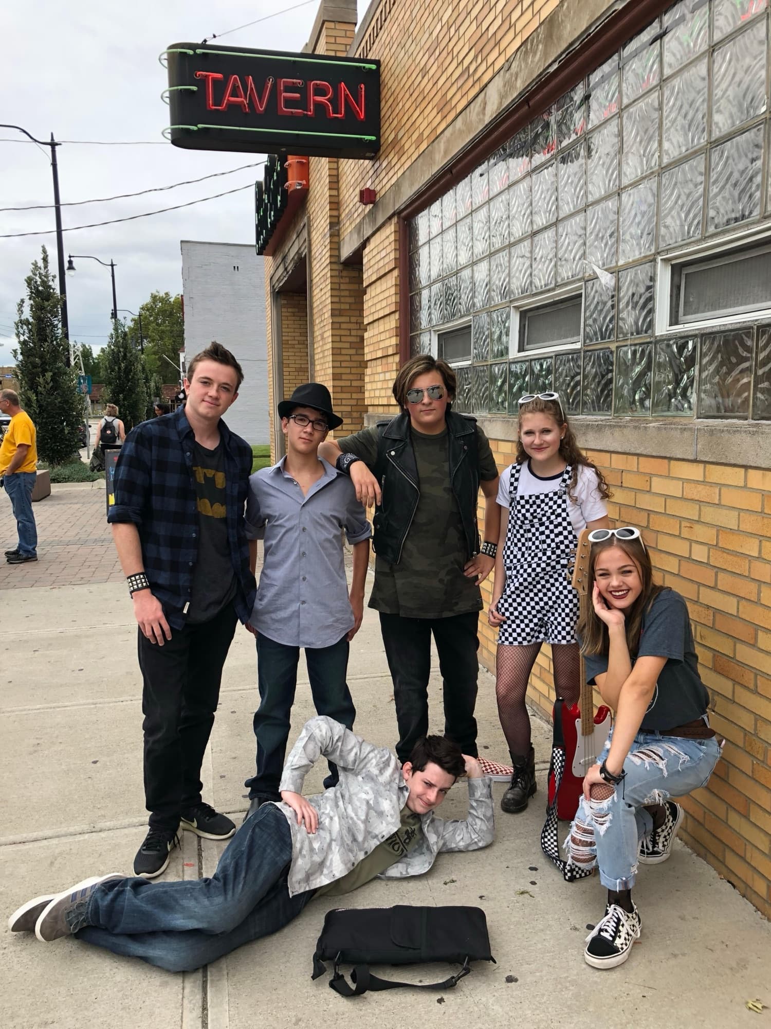 The band posing outside a bar under a neon TAVERN sign, one member sprawled out on the sidewalk in front